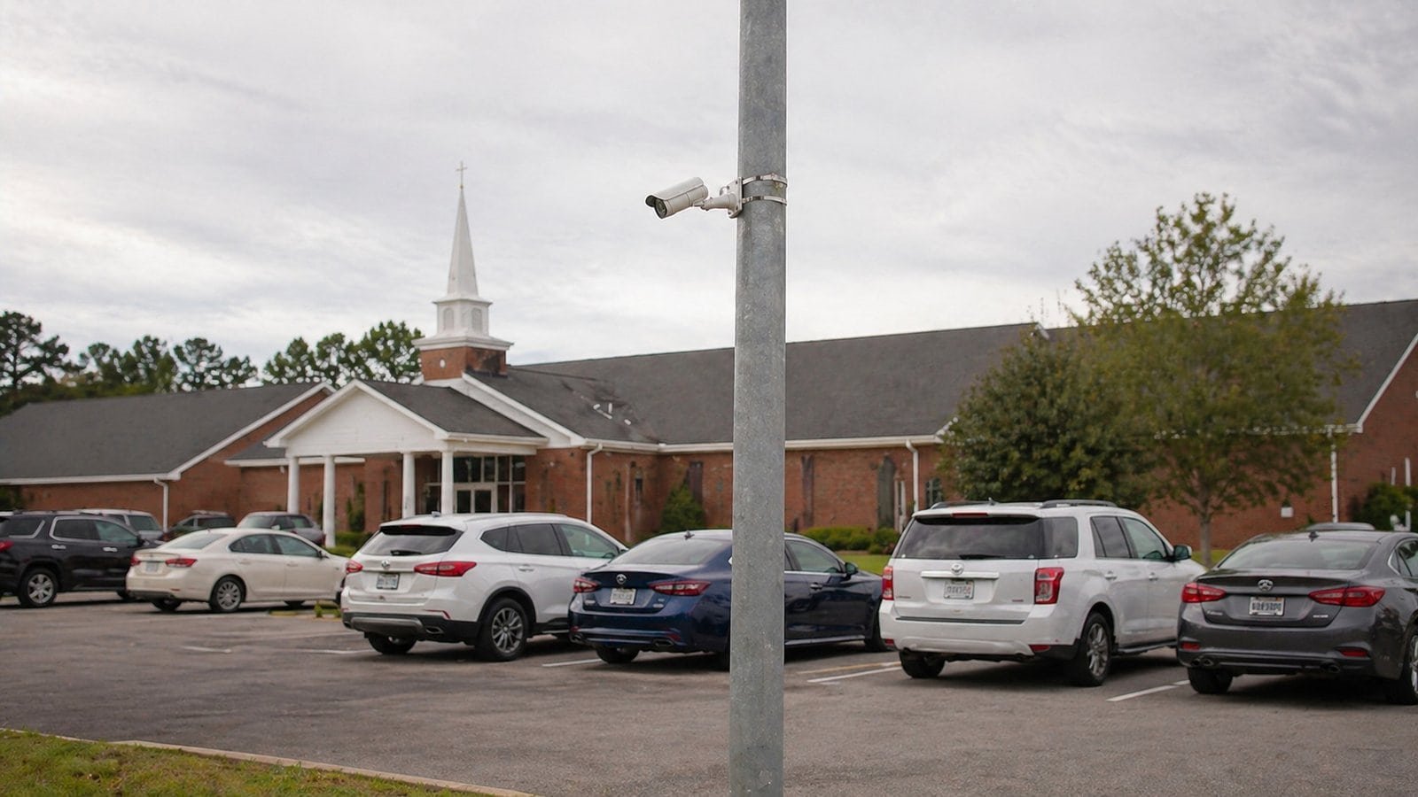 Parking lot with cars in front of a church, featuring a steeple and trees.